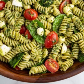 A close-up of a wooden bowl filled with Pesto Pasta Salad—rotini pasta coated in pesto, mixed with cherry tomatoes, mozzarella balls, and fresh spinach leaves.