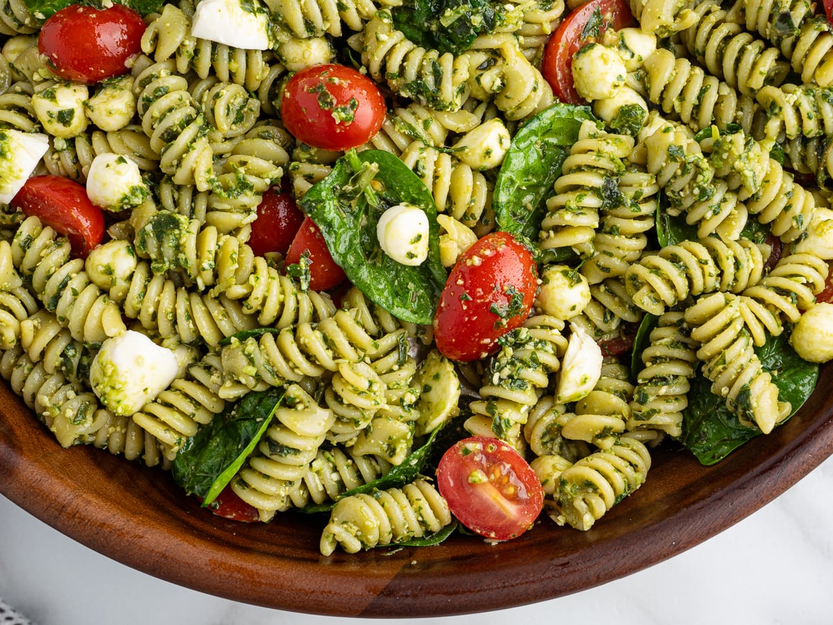 A close-up of a wooden bowl filled with Pesto Pasta Salad—rotini pasta coated in pesto, mixed with cherry tomatoes, mozzarella balls, and fresh spinach leaves.