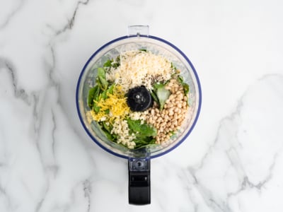 A food processor bowl containing basil leaves, grated parmesan cheese, pine nuts, minced garlic, and lemon zest—perfect ingredients for a fresh Pesto Pasta Salad—sits on a white marble surface.