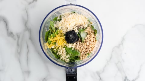 A food processor bowl containing basil leaves, grated parmesan cheese, pine nuts, minced garlic, and lemon zest—perfect ingredients for a fresh Pesto Pasta Salad—sits on a white marble surface.