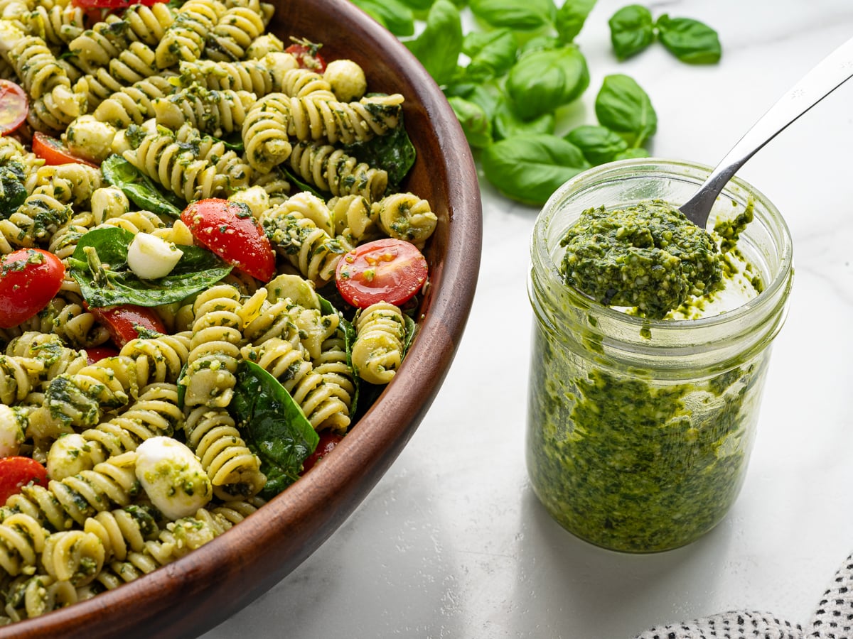 A wooden bowl of Pesto Pasta Salad with rotini, cherry tomatoes, spinach, and mozzarella sits next to a glass jar of green pesto. Fresh basil leaves are scattered on the white surface for a vibrant finish.