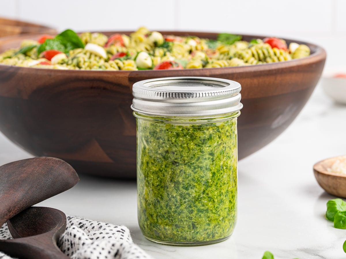 A glass jar filled with green pesto sits on a countertop, in front of a large wooden bowl of Pesto Pasta Salad with tomatoes, mozzarella, and basil. A wooden spoon and a dotted cloth are nearby.