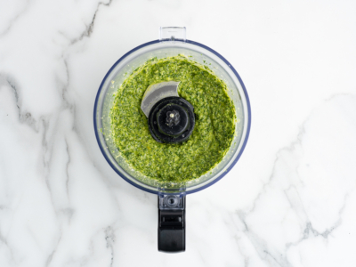 Overhead view of a food processor filled with freshly made green pesto sauce for a vibrant Pesto Pasta Salad, placed on a white marble surface.