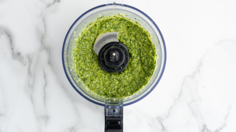 Overhead view of a food processor filled with freshly made green pesto sauce for a vibrant Pesto Pasta Salad, placed on a white marble surface.