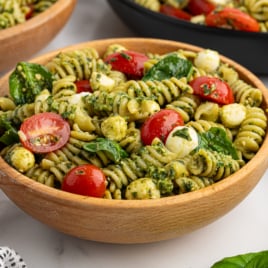 A wooden bowl filled with Pesto Pasta Salad, featuring rotini mixed with cherry tomatoes, fresh spinach, mozzarella balls, and a vibrant pesto sauce.