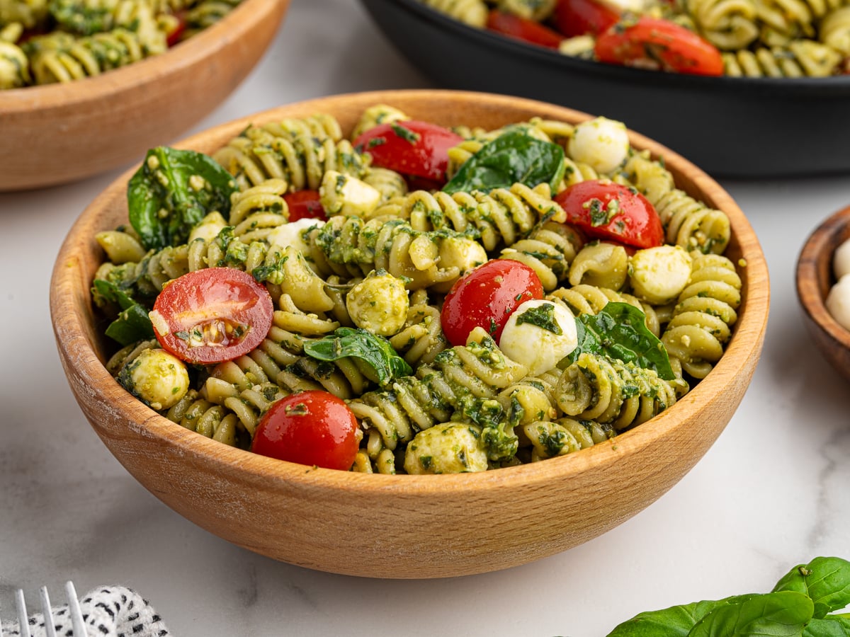 A wooden bowl filled with Pesto Pasta Salad made of rotini noodles, cherry tomatoes, spinach, mozzarella balls, and basil sits on a light surface, with more bowls of pesto pasta salad in the background.