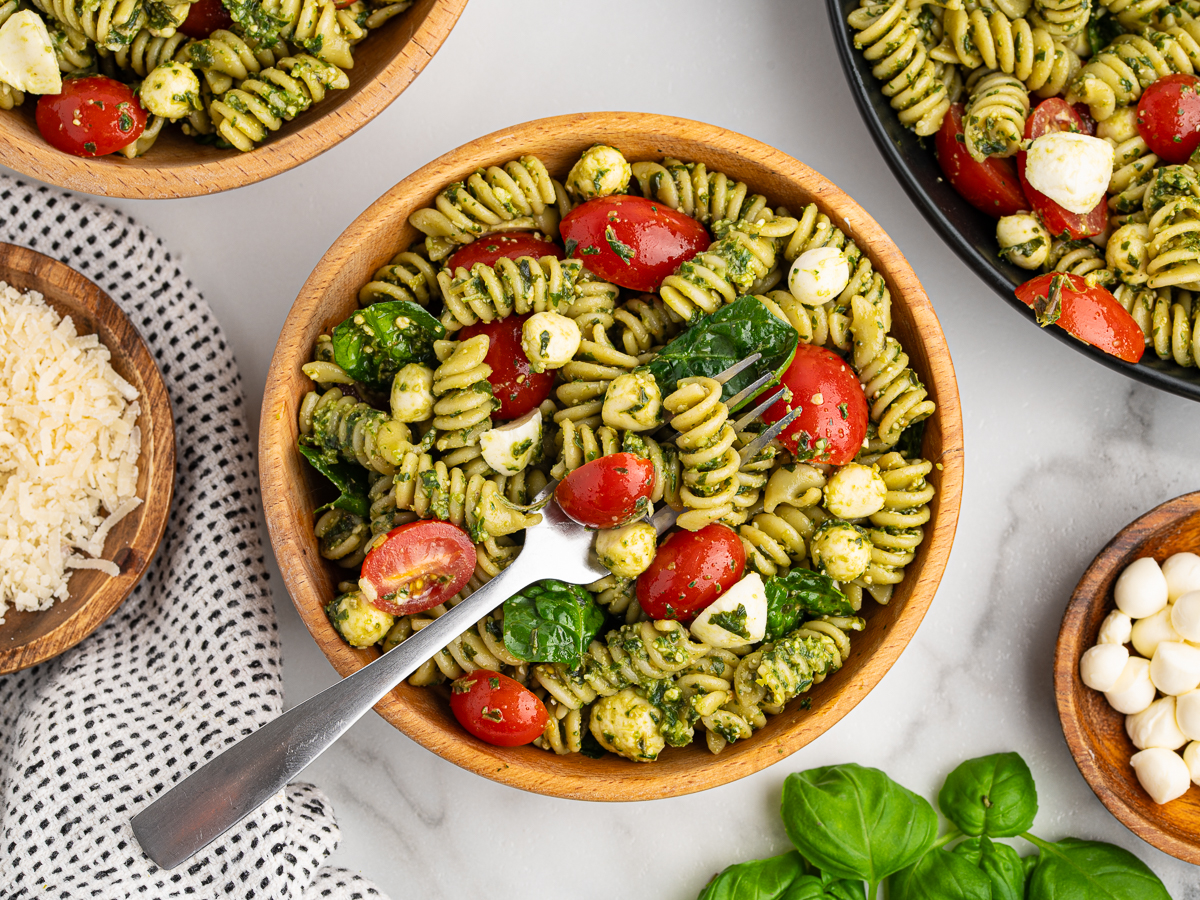 A wooden bowl filled with Pesto Pasta Salad, featuring rotini pasta, cherry tomatoes, mozzarella balls, spinach, and basil. A fork rests in the bowl while nearby bowls of salad, cheese, and fresh basil leaves complete the scene.