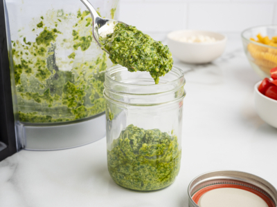 A spoonful of green pesto is being scooped into a glass jar, with a food processor and ingredients for Pesto Pasta Salad arranged on a white countertop in the background.