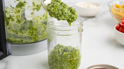 A spoonful of green pesto is being scooped into a glass jar, with a food processor and ingredients for Pesto Pasta Salad arranged on a white countertop in the background.