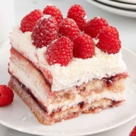 Slice of white chocolate raspberry tiramisu on a white plate, topped with fresh raspberries and white chocolate shavings.