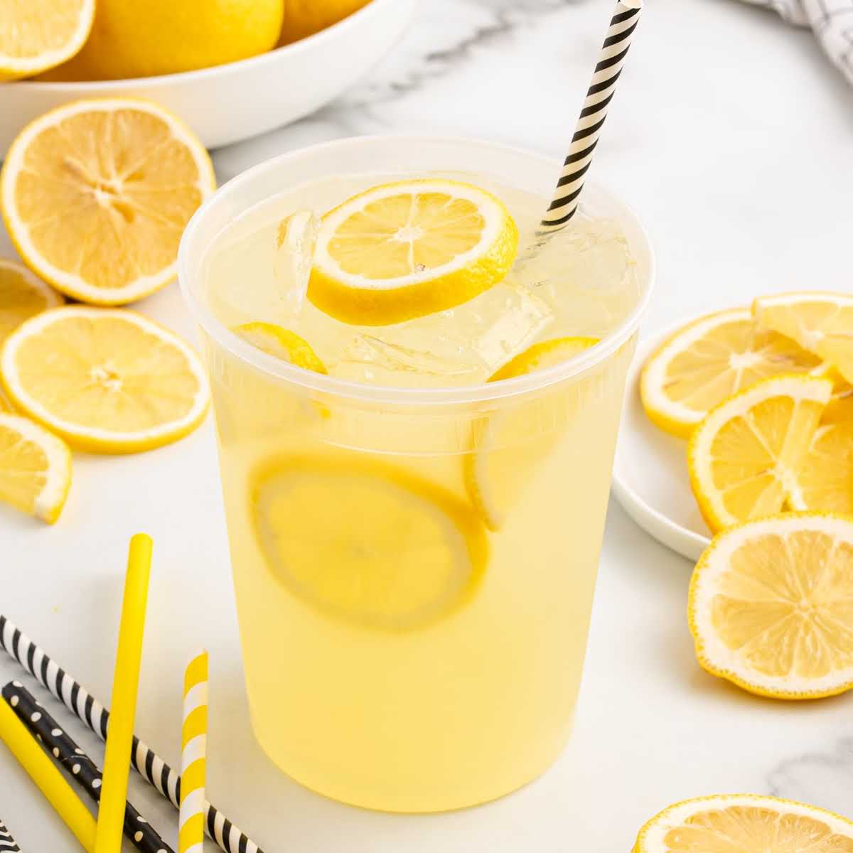 Plastic cup of state fair lemonade with lemon slices and black striped straw surrounded by lemon slices and straws.