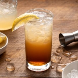 A tall glass of iced cocktail with layered dark and light colors, garnished with a lemon wedge, sits on a wooden surfaceโjust like a perfect Texas Tea Recipeโsurrounded by melting ice, a metal jigger, lemon slices, and a jar of drink.