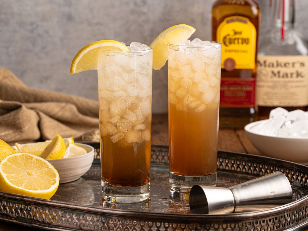 Two tall glasses of iced tea, each with a lemon wedge, sit on a metal tray with a jigger, lemon slices, ice, and bottles of Jose Cuervo and Maker’s Mark—perfect for serving your favorite Texas Tea Recipe.