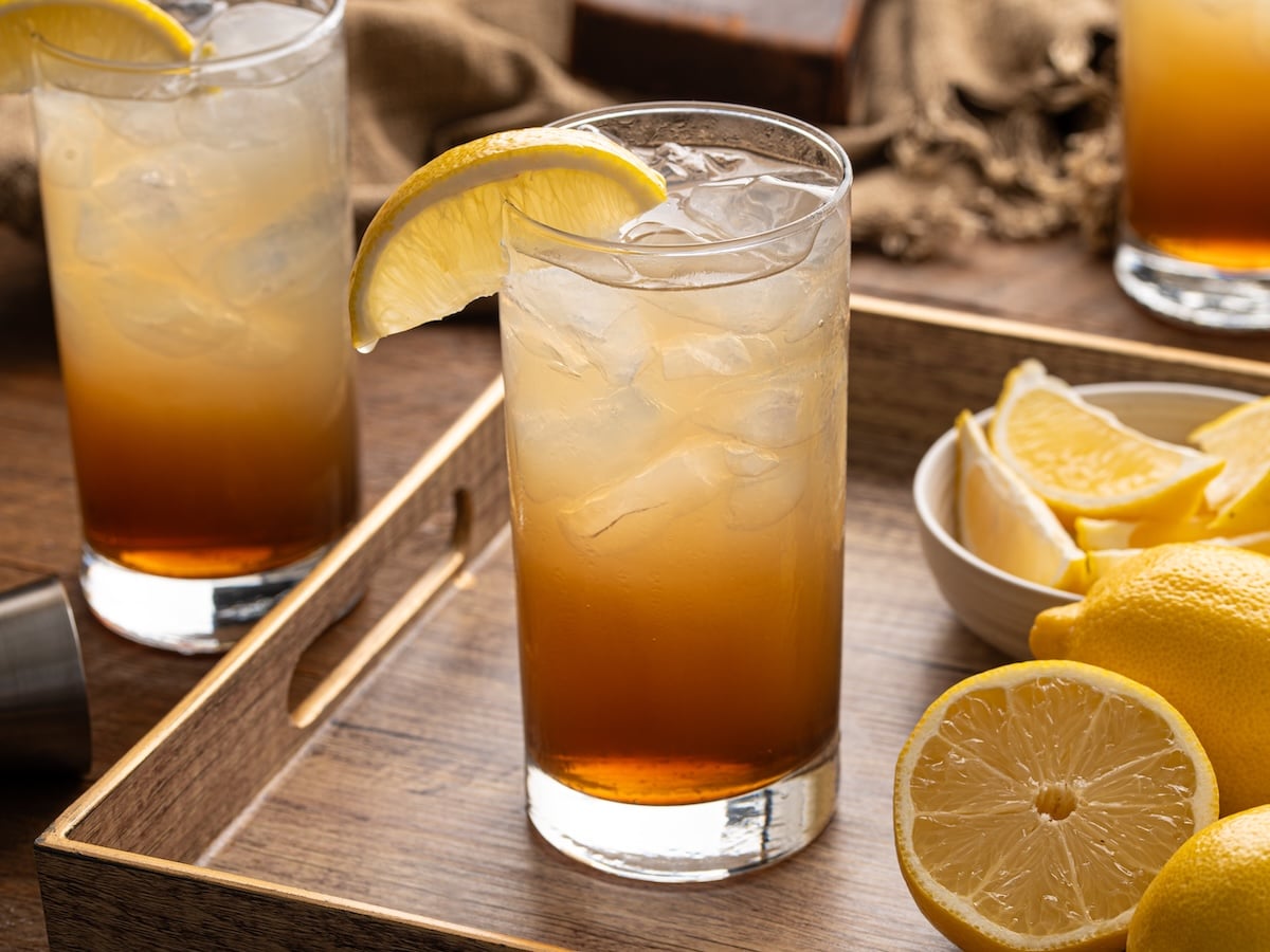 Tall glass of Texas tea with ice and a lemon wedge, showing a layered amber gradient, set on a wooden tray with fresh lemons and a second drink in the background.