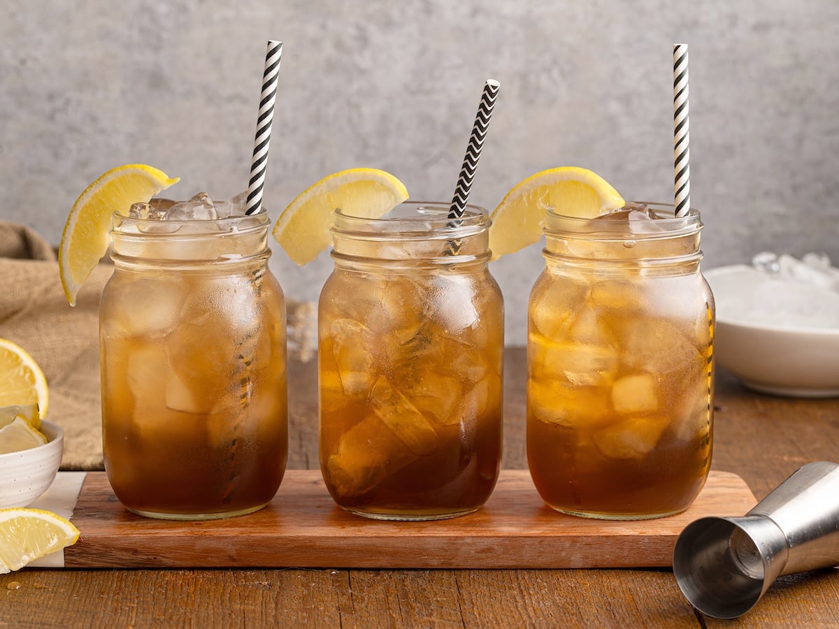 Three mason jars filled with Texas Tea, each garnished with a lemon wedge and a black-and-white striped straw, are arranged on a wooden board. Extra lemon slices and a metal jigger are nearby on the table.