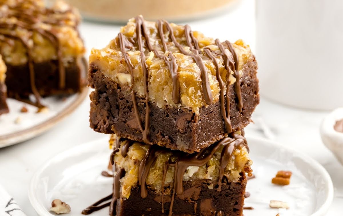 Two thick, fudgy German chocolate brownies stacked on a white plate, topped with coconut-pecan frosting and drizzled with melted chocolate. A blurred background shows more brownies on a plate and a white mug.
