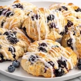Plate of golden-brown blueberry biscuits drizzled with sweet glaze, surrounded by fresh blueberries on a marble surface.