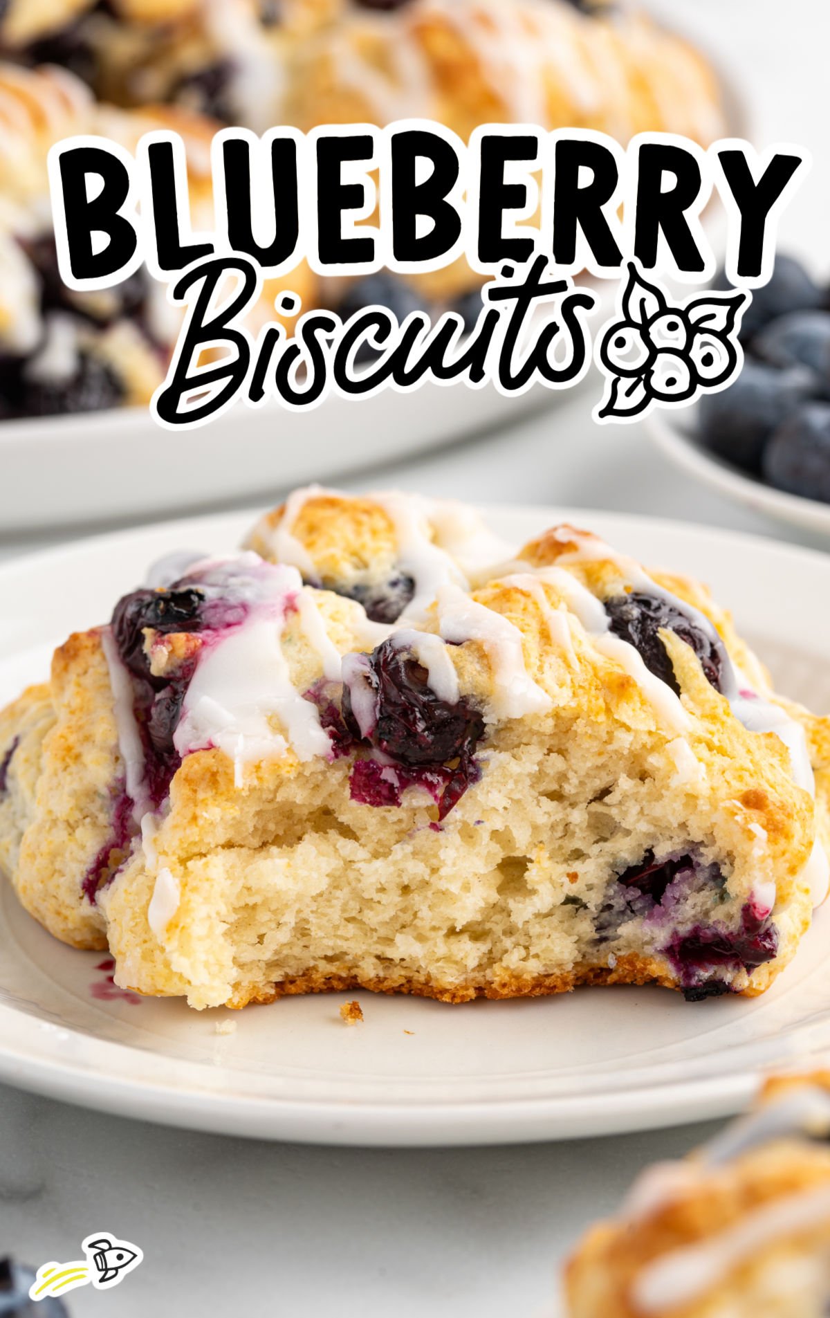 Close-up of a blueberry biscuit on a white plate with a bite taken out, topped with glaze and surrounded by more biscuits in the background.
