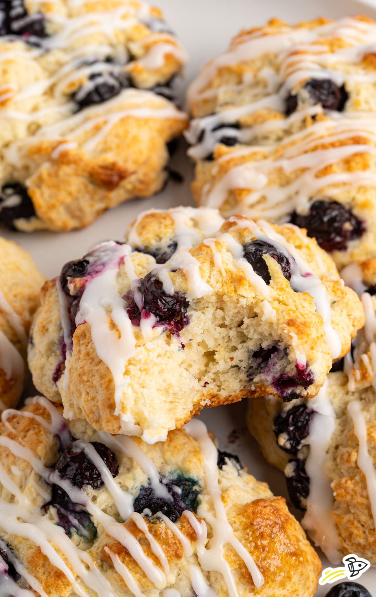Golden blueberry biscuits with a light glaze, one biscuit in the center showing a soft, fluffy inside with juicy blueberries.
