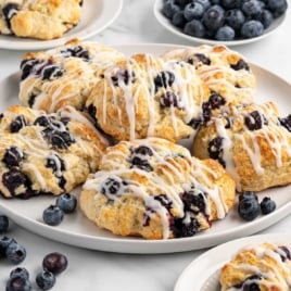 Round white plate filled with glazed blueberry biscuits, surrounded by scattered fresh blueberries and smaller side plates.