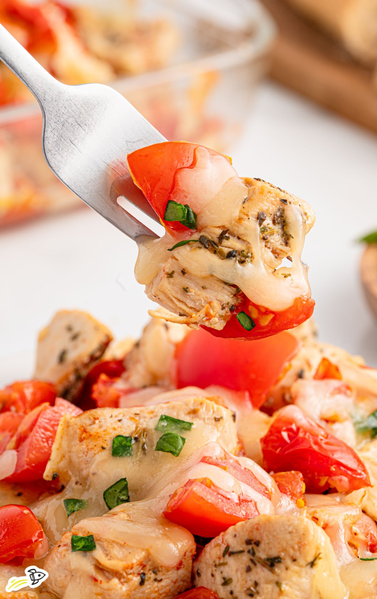 Close-up of a forkful of bruschetta chicken bake with melted mozzarella, juicy tomato, fresh basil, and seasoned seared chicken, lifted from a serving plate.