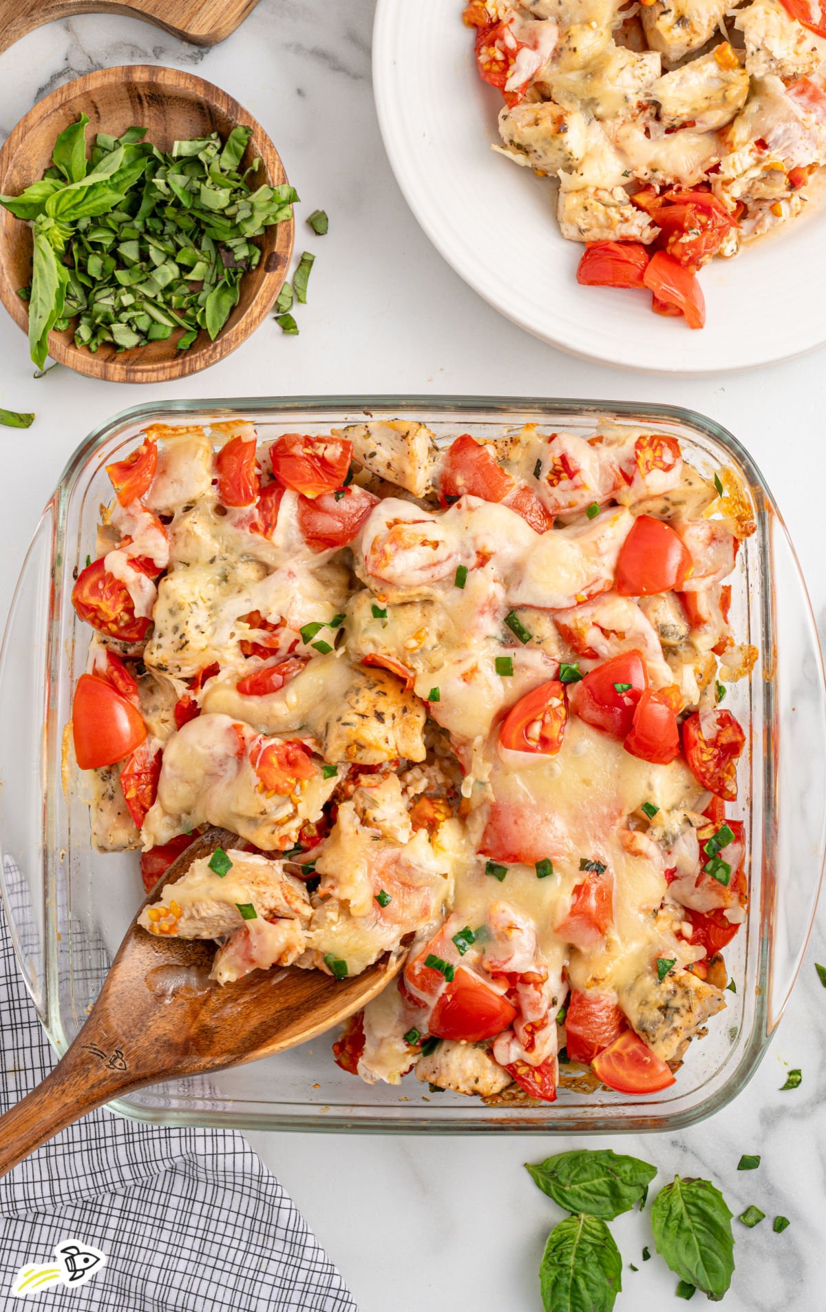 Fully baked bruschetta chicken bake in a glass dish with melted mozzarella, juicy tomatoes, and chopped basil, with a wooden spoon serving a portion.
