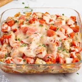 Glass baking dish filled with bruschetta chicken bake, surrounded by fresh basil, plates, and forks on a marble countertop.