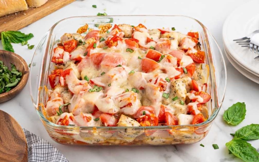 Glass baking dish filled with bruschetta chicken bake, surrounded by fresh basil, plates, and forks on a marble countertop.