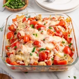 Top-down view of baked bruschetta chicken in a glass dish, garnished with chopped basil, next to a bowl of fresh herbs and stack of white plates.