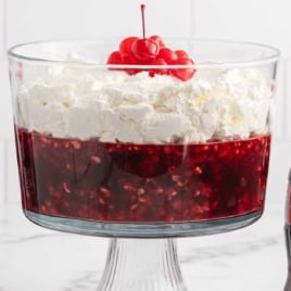 Glass trifle bowl filled with Coca Cola fruit trifle topped with whipped cream and cherries, surrounded by cherry pie filling, Jell-O box, and a glass Coke bottle.