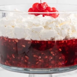 Glass trifle bowl filled with Coca Cola fruit trifle topped with whipped cream and cherries, surrounded by cherry pie filling, Jell-O box, and a glass Coke bottle.