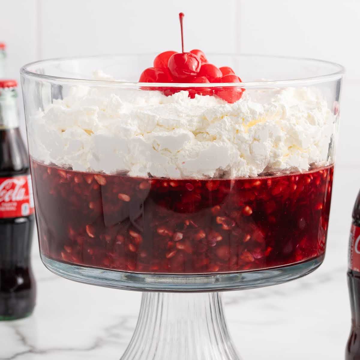 Glass trifle bowl filled with Coca Cola fruit trifle topped with whipped cream and cherries, surrounded by cherry pie filling, Jell-O box, and a glass Coke bottle.