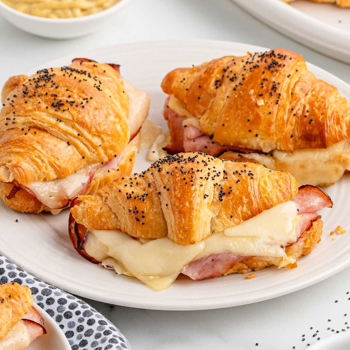 Plate of baked ham and cheese croissants with melted cheese and sprinkled poppy seeds, served with a bowl of mustard sauce