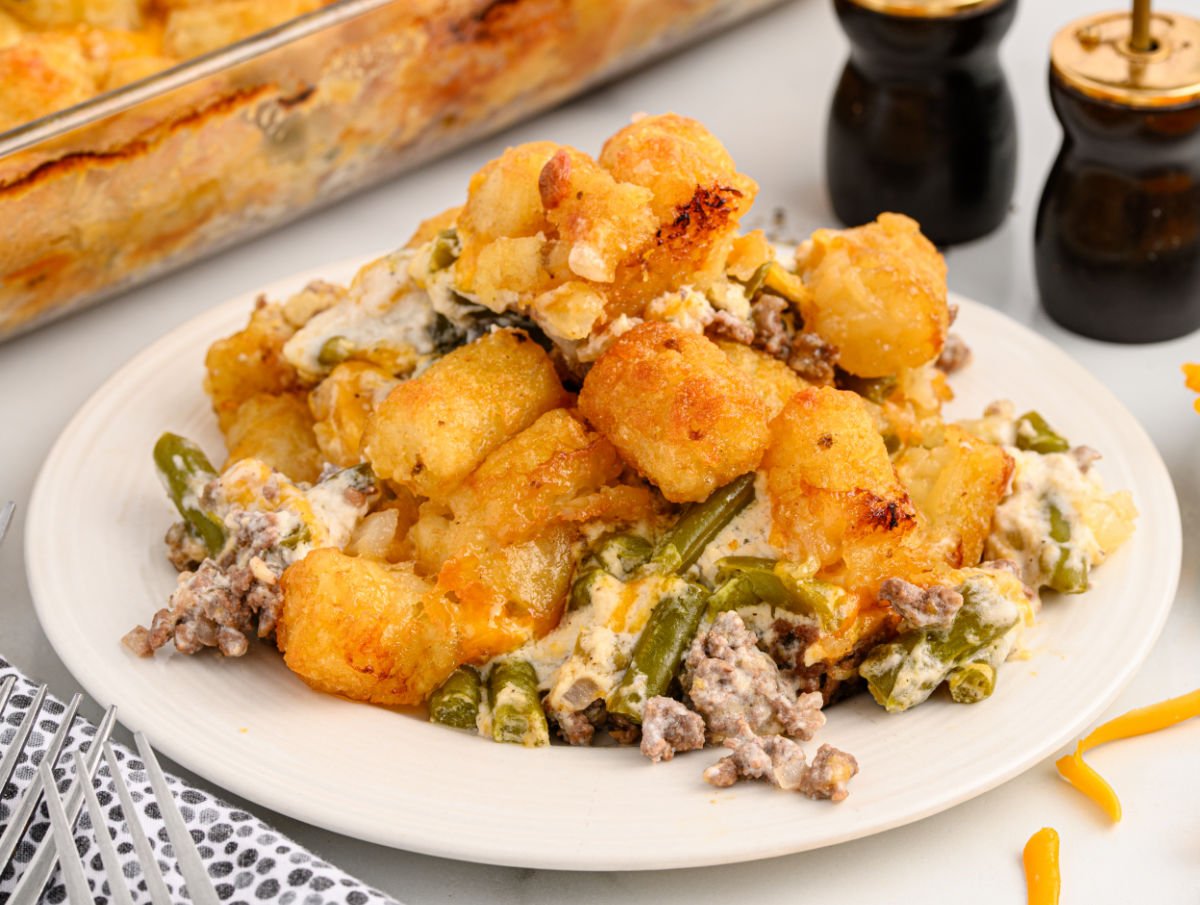 A serving of hamburger tater tot casserole on a white plate, showing layers of golden, crispy tater tots, creamy sauce, ground beef, and green beans, with the full casserole dish in the background.