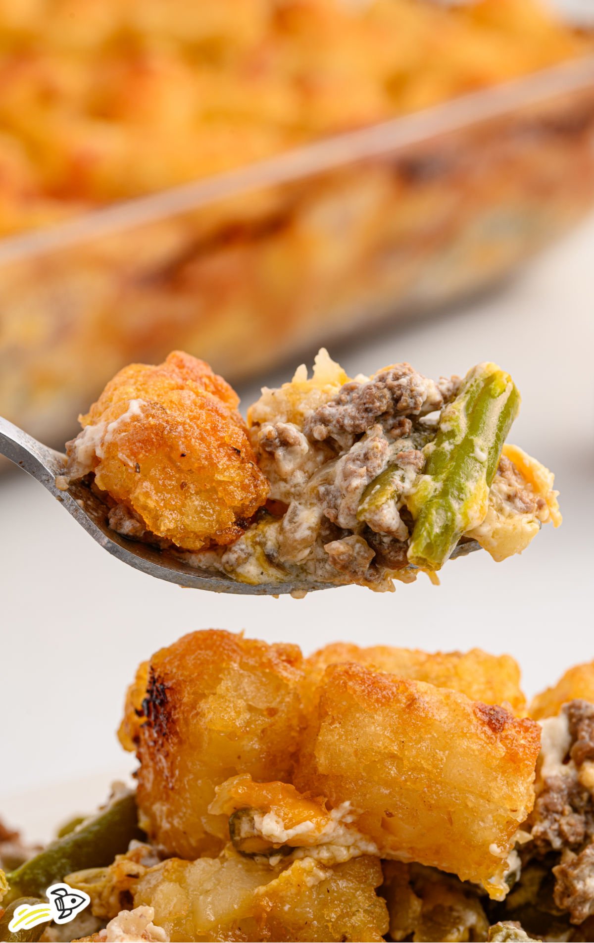A close-up of a spoonful of hamburger tater tot casserole, highlighting the crunchy tater tots, seasoned ground beef, creamy sauce, and tender green beans, with more casserole visible in the background.