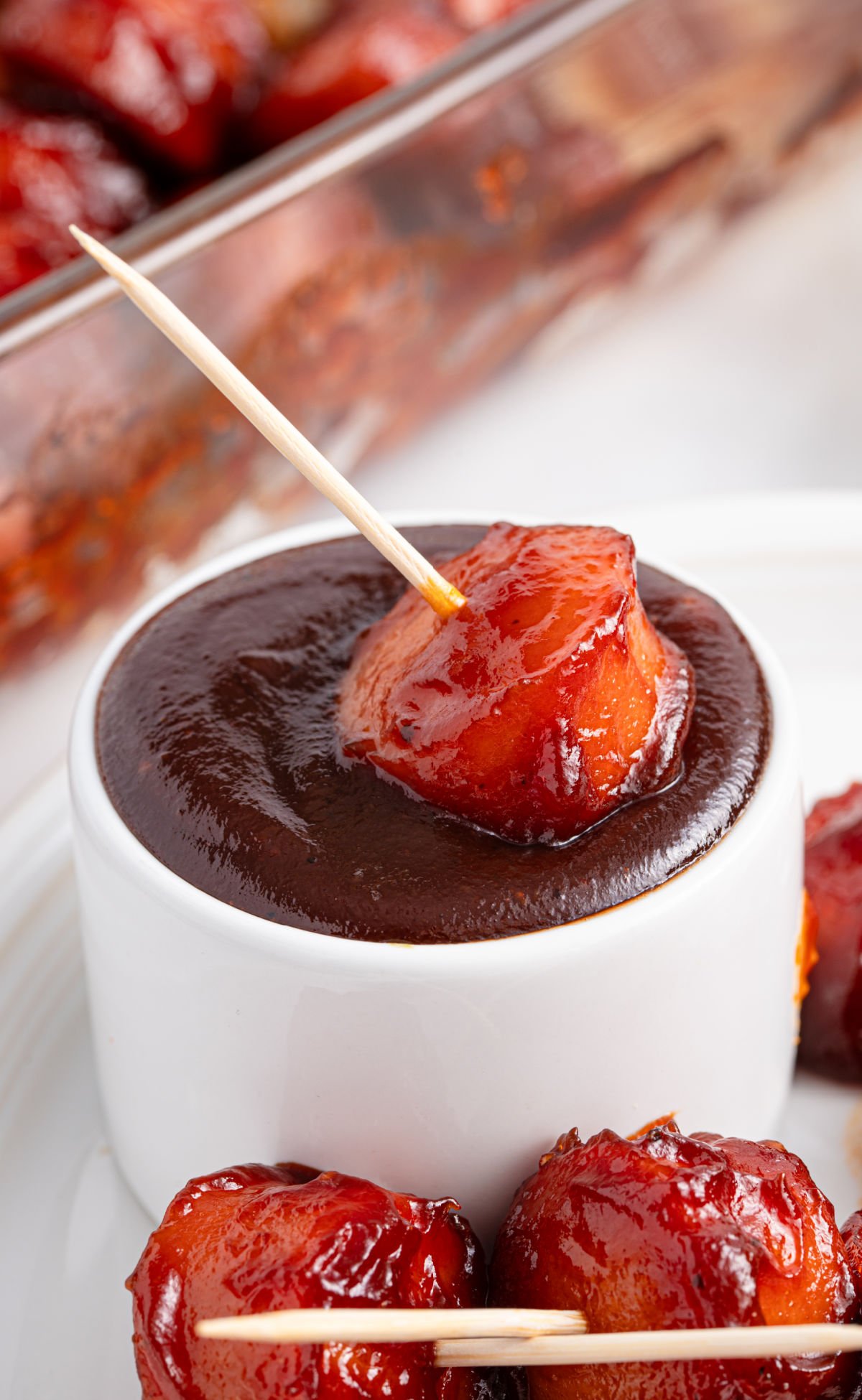 Close-up of a glazed BBQ sausage bite on a toothpick being dipped into a rich, dark barbecue sauce in a white ramekin.