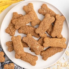 Plate of homemade banana dog treats shaped like bones surrounded by bananas, oats, and a bowl of peanut butter.