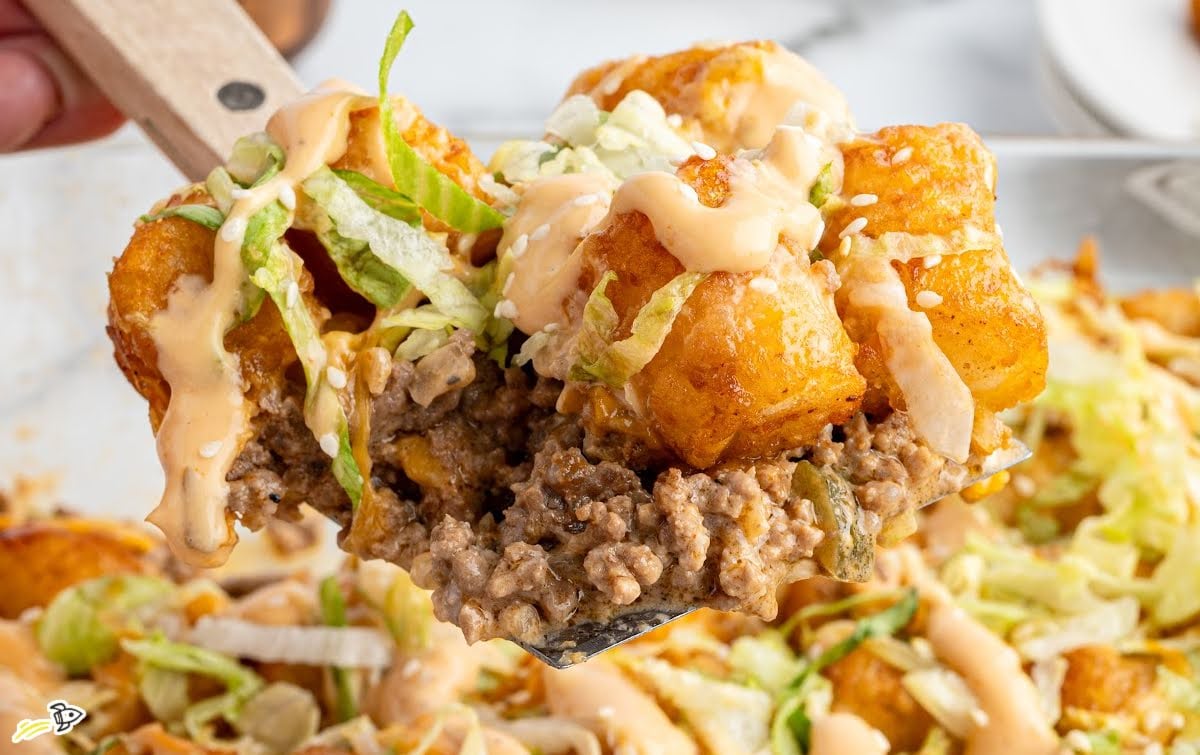 Serving spoon lifting a portion of Big Mac tater tot casserole with ground beef, tater tots, lettuce, and sauce.