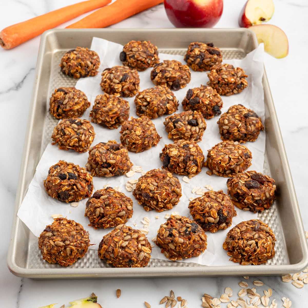 Full tray of baked goat treats on parchment paper with fresh carrots and apples beside it.