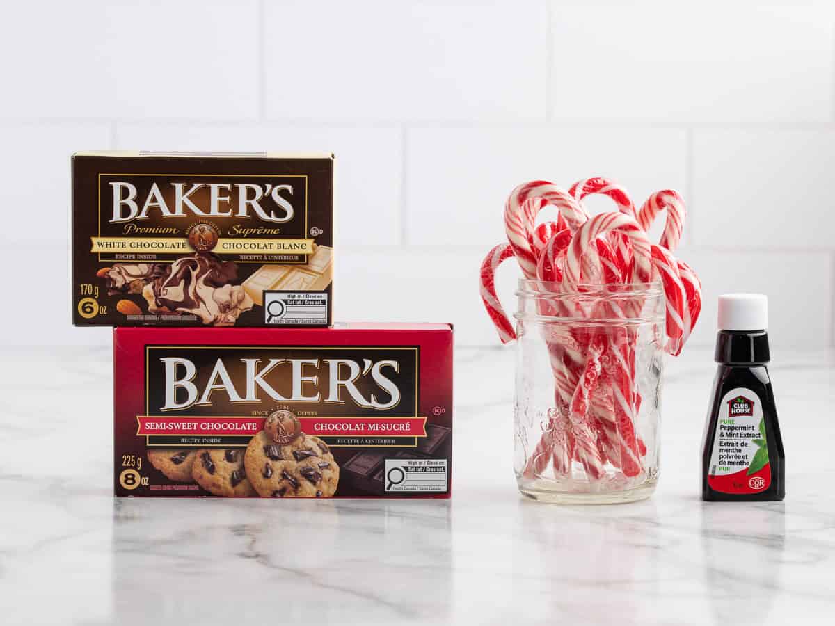 Two boxes of Bakers chocolate, a glass jar filled with mini candy canes, and a small bottle of peppermint extract sit on a marble counter with a white tile background—the perfect setup for making your favorite Peppermint Bark Recipe.