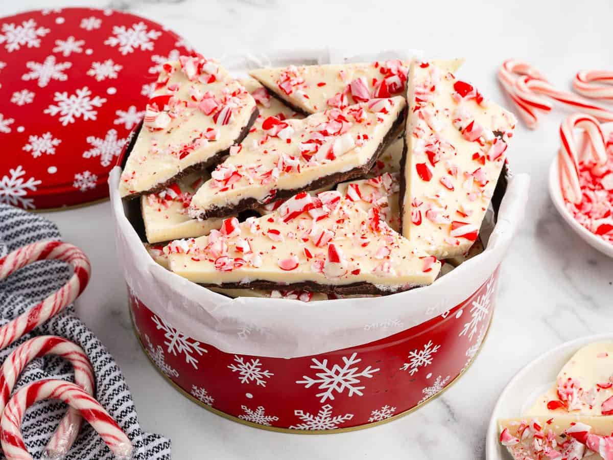 A festive tin with white snowflakes holds pieces of peppermint bark crafted from a classic Peppermint Bark Recipe, each topped with crushed candy canes. Candy canes and a black-and-white striped cloth are nearby.