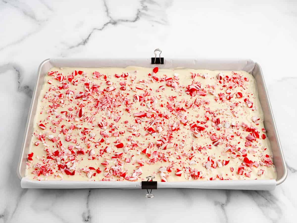 A baking sheet lined with parchment paper holds Peppermint Bark topped with crushed red and white candy canes, set on a marble surface.