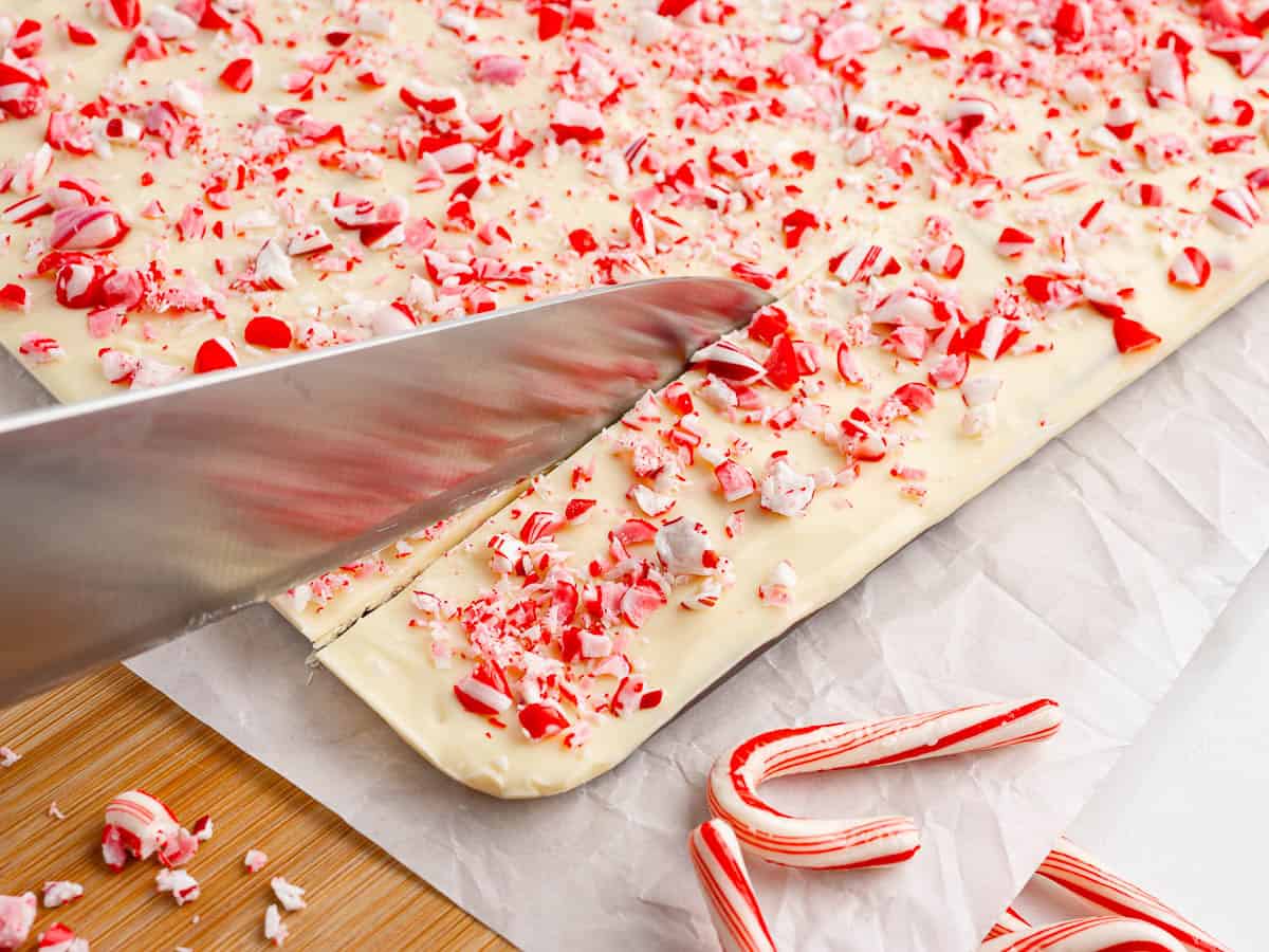 A large knife cuts a slab of peppermint bark—creamy white chocolate topped with crushed peppermint candies—while whole candy canes rest nearby on parchment paper, ready to complete this classic Peppermint Bark Recipe.