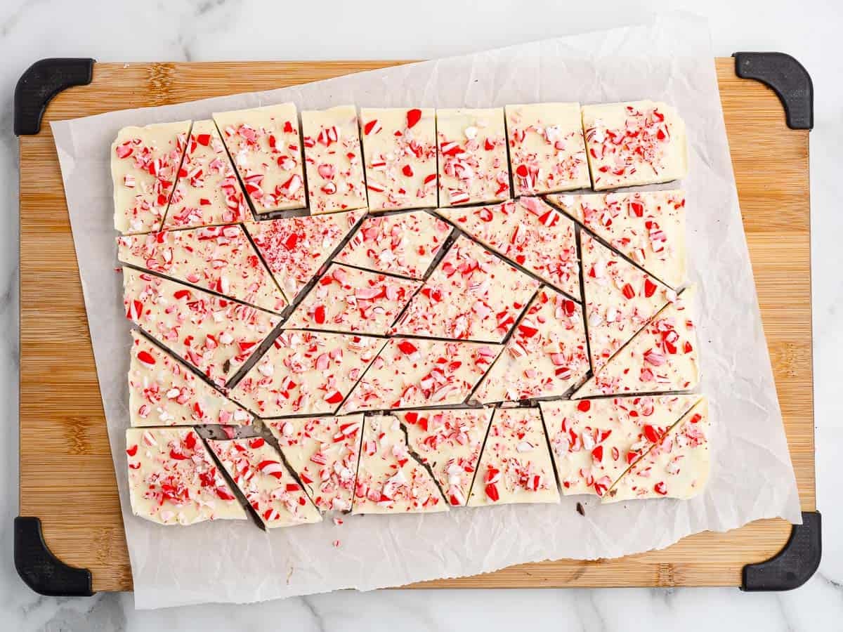 White chocolate peppermint bark, made using a classic Peppermint Bark Recipe, is broken into pieces on parchment paper atop a wooden cutting board, with red and white candy bits sprinkled on top.