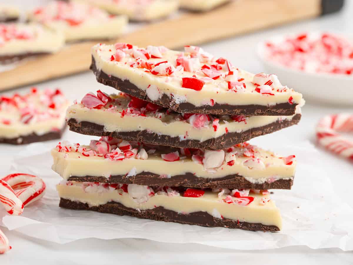 Three stacked pieces of Peppermint Bark with a dark chocolate base, white chocolate layer, and crushed candy canes on top, sitting on parchment paper. More bark and candy canes are visible in the background, creating an irresistible scene that’s perfect inspiration for your next Peppermint Bark recipe.