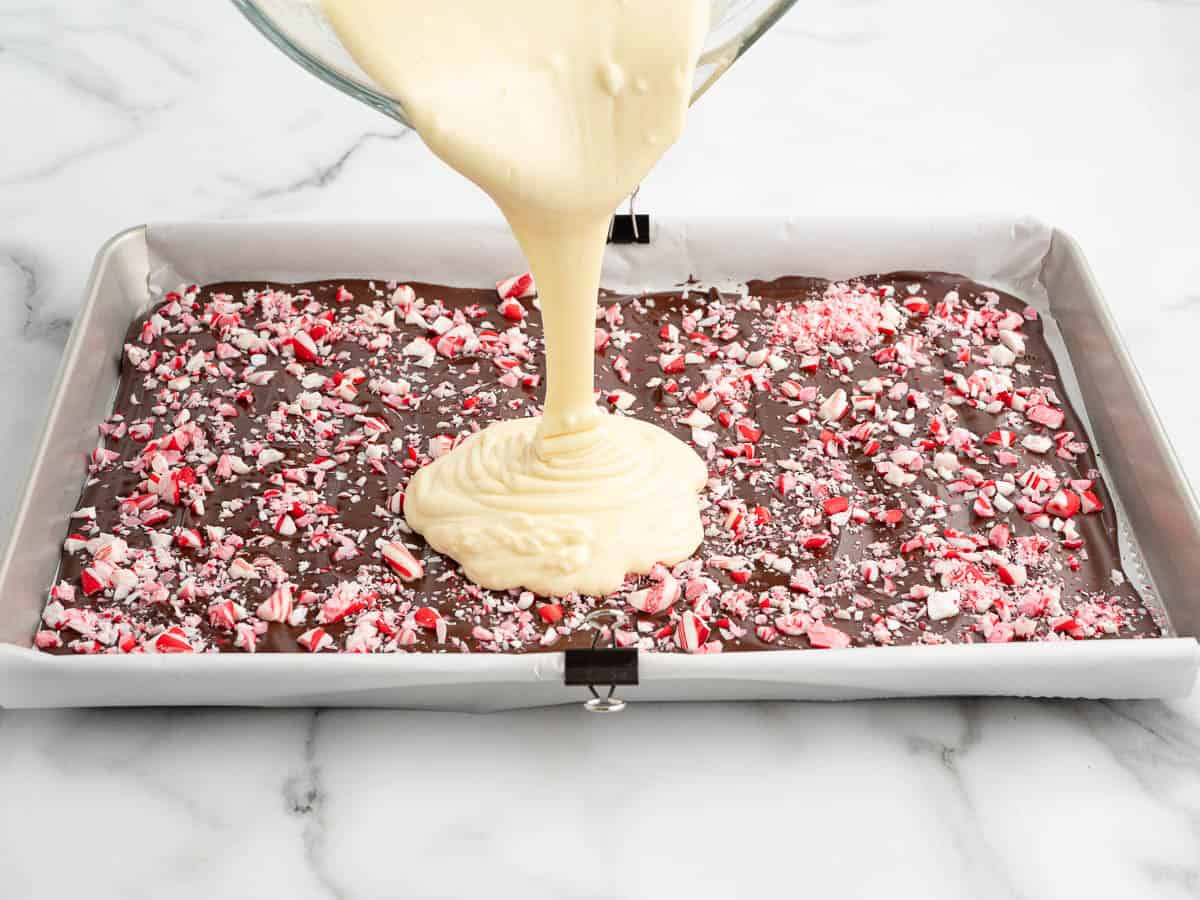 White chocolate is being poured over a layer of dark chocolate topped with crushed peppermint candies on a parchment-lined baking sheet—creating the perfect Peppermint Bark Recipe.