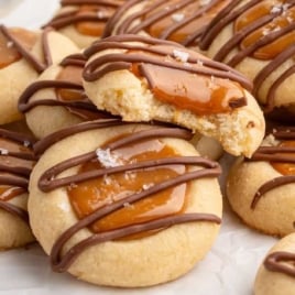 Pile of Twix thumbprint cookies topped with melted caramel, chocolate drizzle, and flaky sea salt