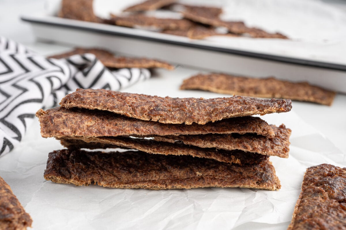 A stack of thin, rectangular, dark brown crispbread slices sits on white parchment paper, resembling the wholesome look of Beef Jerky for Dogs, with more crispbread and a black-and-white patterned cloth in the background.