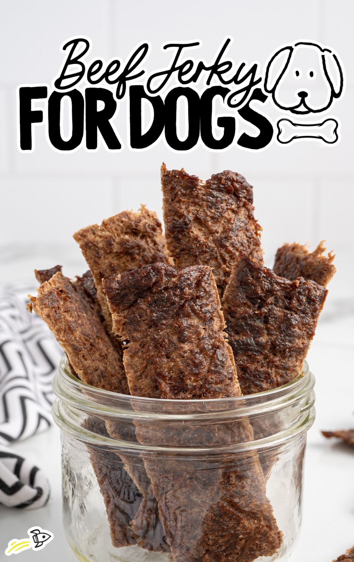a jar filled with homemade beef jerky strips for dogs on a white surface with a black and white patterned towel.