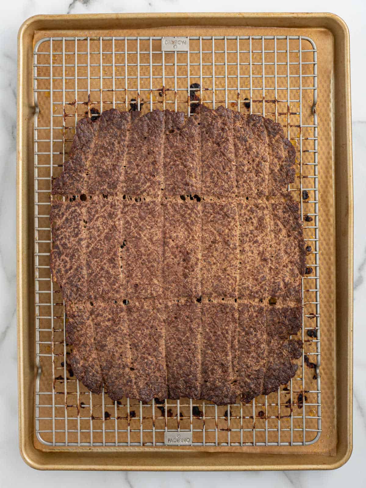 A large sheet of browned beef jerky is cooling on a wire rack over a baking sheet, divided into rectangular sections with visible score marks—perfectly prepared as homemade beef jerky for dogs.